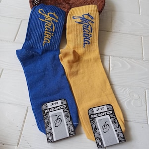 Puede incluir: Un par de calcetines, uno azul y otro amarillo, con la palabra "Ukraina" escrita en azul en cada calcetín. Los calcetines están etiquetados con "UKRAPETKIN" y "25-27".