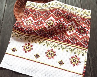 Puede incluir: Toalla de cocina blanca con un patrón geométrico bordado en rojo, amarillo y negro. La toalla está doblada y colocada sobre una superficie de madera.