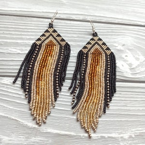 May include: A pair of long, beaded earrings with a geometric pattern in black, gold, and brown. The earrings are made of small beads and have a fringe-like design.