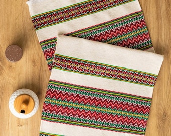 Puede incluir: Dos caminos de mesa blancos con patrones bordados en rojo, verde y amarillo. Los patrones son geométricos e incluyen zigzags y diamantes.