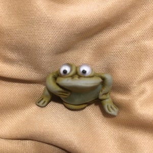 May include: A small green ceramic frog figurine with large white eyes. The frog is sitting with its arms crossed and its legs bent.