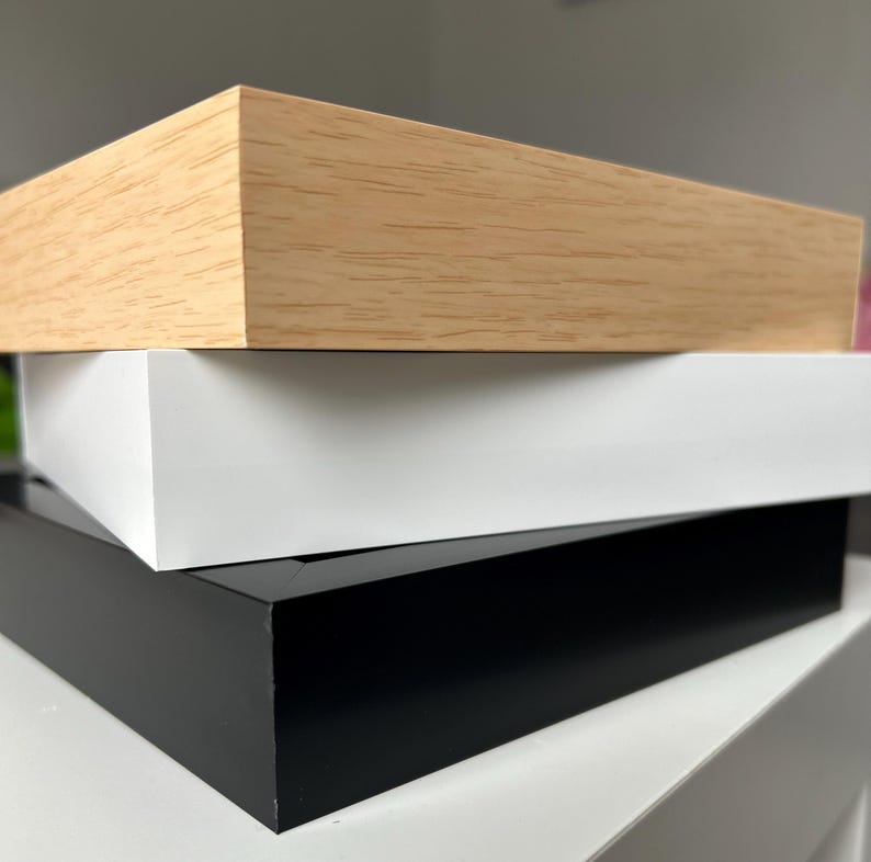 May include: Three picture frames stacked on top of each other. The top frame is made of light wood, the middle frame is white, and the bottom frame is black.