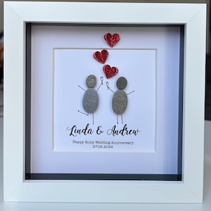 May include: A framed artwork featuring two pebble figures holding up glasses, with three red glitter hearts above them. The text below reads "Linda & Andrew Happy Ruby Wedding Anniversary 27.06.2024".