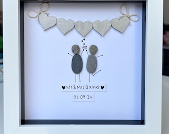 May include: A white frame with a white mat and a black background. The frame contains a pebble art picture of a couple toasting with a string of gray hearts above them. The text below the couple reads "Mr. & Mrs. Garner 21.09.24".
