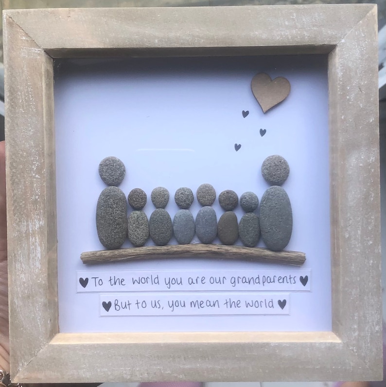 K&ouml;nnte beinhalten: Ein gerahmtes Kunstwerk aus Kieselsteinen mit der Aufschrift "F&uuml;r die Welt seid ihr unsere Gro&szlig;eltern, aber f&uuml;r uns seid ihr die Welt". Das Kunstwerk zeigt eine Familie aus sieben Personen, die durch Kieselsteine dargestellt werden und auf einem St&uuml;ck Treibholz sitzen. &Uuml;ber der Familie schweben kleine braune Papierherzen.