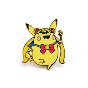 May include: A gold-colored enamel pin featuring a yellow cartoon character with a pink bow, blue sailor collar, and a crescent moon headband. The character is holding a wand with a crescent moon on top.