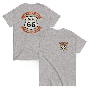 Puede incluir: Dos camisetas grises con gráficos de la Ruta 66. Una camiseta presenta un diseño grande en la espalda con el texto "The Mother Road" y "U.S. Route 66 100th Anniversary". La otra camiseta tiene un logotipo de la Ruta 66 más pequeño en la parte delantera.