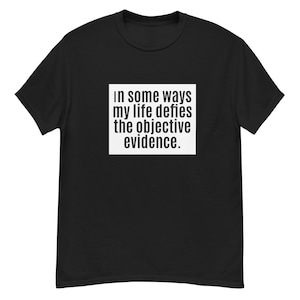 May include: Black t-shirt with white text that reads "In some ways my life defies the objective evidence."