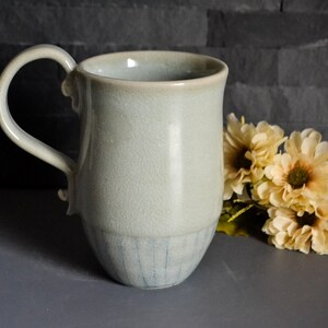 Könnte beinhalten: Handgefertigte Keramiktasse mit hellblauer Glasur. Die Tasse hat einen großen Henkel und eine strukturierte Oberfläche. Der Boden der Tasse weist vertikale Linien auf. Dekorative Blumen befinden sich im Hintergrund.