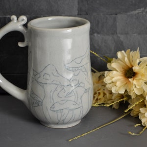 Pode incluir: Uma caneca de cer&acirc;mica com um esmalte cinza claro e uma al&ccedil;a &uacute;nica. A caneca apresenta um desenho de cogumelos desenhado &agrave; m&atilde;o em preto.