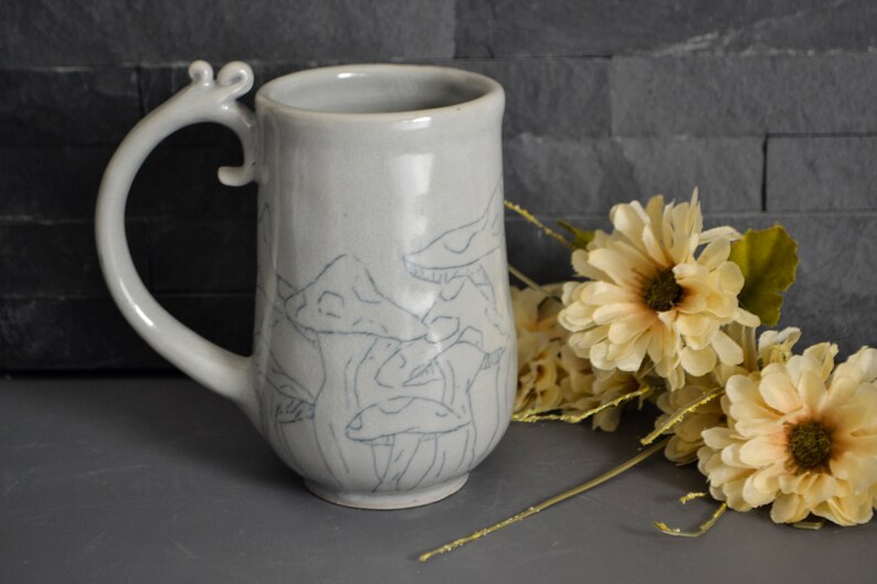 Pode incluir: Uma caneca de cer&acirc;mica com um esmalte cinza claro e uma al&ccedil;a &uacute;nica. A caneca apresenta um desenho de cogumelos desenhado &agrave; m&atilde;o em preto.