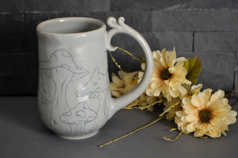 Pode incluir: Uma caneca de cer&acirc;mica cinza com uma al&ccedil;a &uacute;nica e um desenho de cogumelos em uma cor cinza claro.