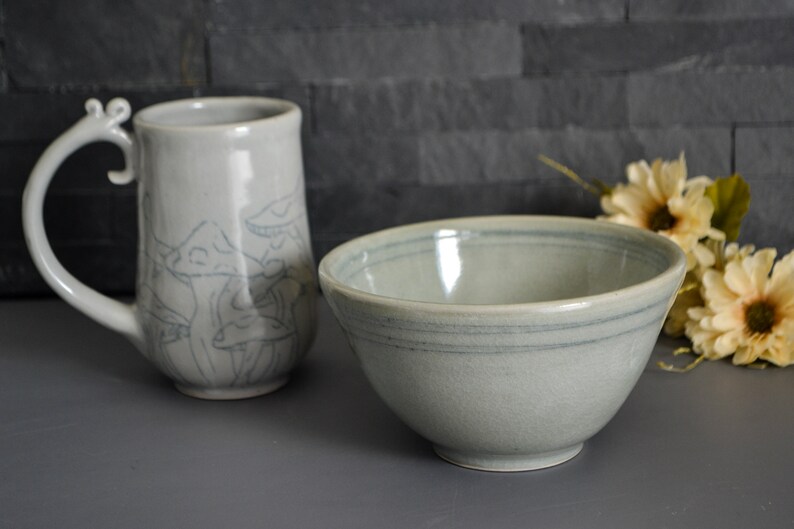 Pode incluir: Uma caneca de cer&acirc;mica branca com al&ccedil;a e um desenho de cogumelos, e uma tigela de cer&acirc;mica cinza claro com um design de listras azuis.