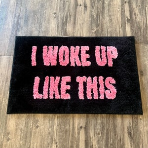 Puede incluir: Felpudo negro con el texto "I woke up like this" en letras rosas esponjosas.