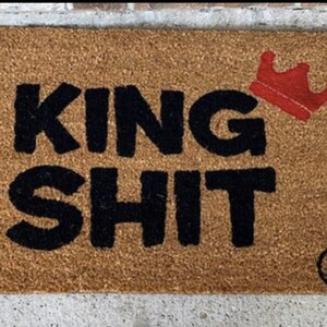 May include: A brown coir doormat with the text "KING SHIT" in black lettering. A red crown is positioned above the text. The doormat has a small black circle with a white "M" in the bottom right corner.