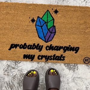 以下が含まれることがあります： 「probably charging my crystals」という文字とカラフルなクリスタル柄が描かれた茶色の玄関マット。マットの右下には小さな黒い「M」のロゴがある。