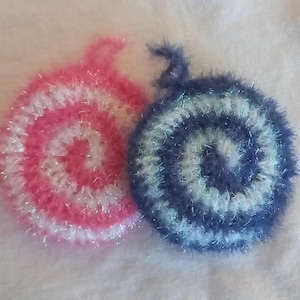 May include: Two round, crocheted scrubbers with a spiral pattern. One scrubber features alternating pink and white fuzzy yarn, while the other features alternating dark blue and white fuzzy yarn. Each scrubber includes a small loop at the top.