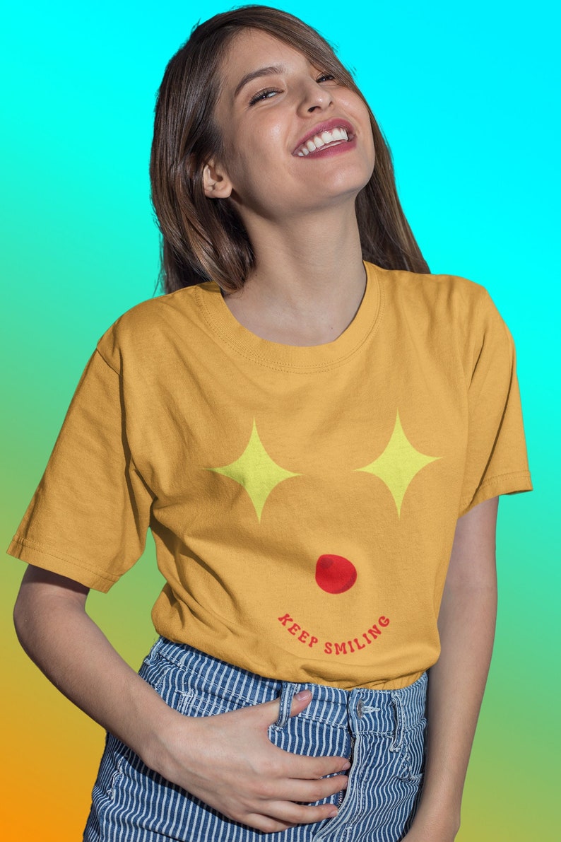 Puede incluir: Una mujer con una camiseta amarilla mostaza con un gr&aacute;fico de una cara sonriente y el texto "KEEP SMILING". Lleva pantalones vaqueros de rayas azules y blancas y tiene el pelo largo y casta&ntilde;o.