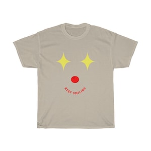 Puede incluir: Una camiseta gris claro con un dise&ntilde;o de cara sonriente. La cara tiene dos estrellas amarillas para los ojos, un c&iacute;rculo rojo para la nariz y el texto "KEEP SMILING" en rojo.