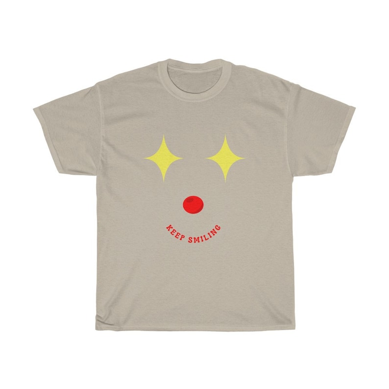 Puede incluir: Una camiseta gris claro con un dise&ntilde;o de cara sonriente. La cara tiene dos estrellas amarillas para los ojos, un c&iacute;rculo rojo para la nariz y el texto "KEEP SMILING" en rojo.