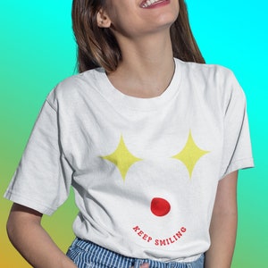Puede incluir: Una camiseta blanca con un gr&aacute;fico de cara sonriente amarilla y el texto "KEEP SMILING". La modelo lleva pantalones a rayas azules y blancas y sonr&iacute;e.