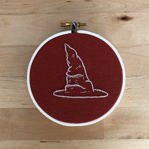 May include: A white embroidered image of a wizard's hat on a red fabric background. The hat is a classic pointed wizard's hat with a brim.
