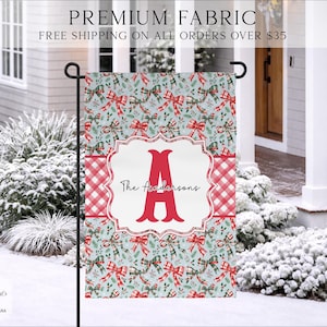 May include: A rectangular garden flag featuring a light blue background with a repeating pattern of red and green plaid bows and holly leaves. A central white scalloped frame contains a large red letter A and the text The Andersons.
