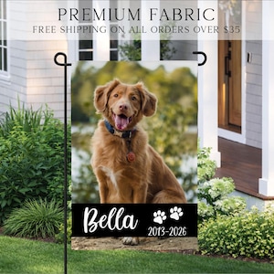 May include: A rectangular garden flag featuring a photographic portrait of a golden-brown dog wearing a blue collar. Below the dog, a black horizontal band displays the name Bella in white script, two white paw prints, and the years 2013-2026.