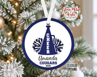 May include: A round white Christmas ornament with a dark blue border and a white ribbon hanger. The design features a dark blue megaphone with the word CHEER, two pom-poms, the name Amanda, the team name COUGARS, and the year 2026.