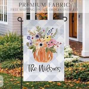 May include: A rectangular garden flag featuring a light blue and white buffalo check pattern. The center displays a watercolor illustration of a pumpkin filled with colorful wildflowers. The text "The Wilsons" is printed in black script below.