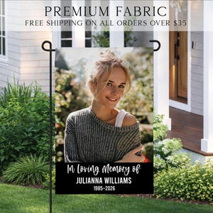 May include: A rectangular garden flag featuring a photographic portrait of a person with blonde hair wearing a grey sweater. Below the image, a black horizontal band displays the white text: In Loving Memory of JULIANNA WILLIAMS 1985-2026.