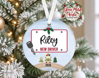 May include: A round, light blue Christmas ornament featuring a white license plate graphic with the name Riley and the text NEW DRIVER. It includes a car key illustration, a small car with gifts, two pine trees, and the year 2026 on a road.