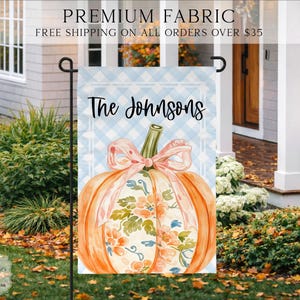 May include: A rectangular garden flag featuring a light blue and white buffalo check background. The center displays an orange pumpkin with a floral pattern and a pink ribbon bow. The text "The Johnsons" is printed in black script at the top.