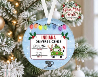 May include: A round, light blue Christmas ornament featuring a graphic of an Indiana driver's license with the name Danielle and the year 2026. The design includes a green truck with a reindeer and Santa, colorful string lights, and car keys.
