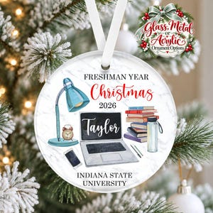 May include: A round white marble-patterned Christmas ornament featuring a blue desk lamp, a laptop with the name Taylor, a stack of books, a water bottle, and a clock. Text reads Freshman Year Christmas 2026 and Indiana State University.