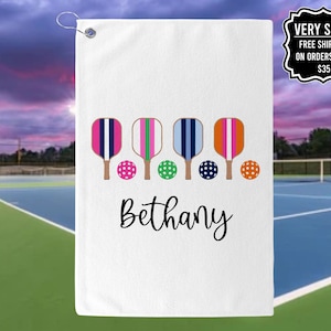 May include: White towel with a personalized name "Bethany" printed in black cursive script. The towel features five colorful paddleball rackets and balls in a row.