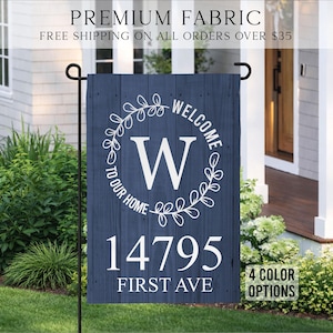 May include: A rectangular garden flag with a dark blue wood grain background. The white text reads WELCOME TO OUR HOME, the letter W, 14795, and FIRST AVE. A white circular vine graphic surrounds the letter W on this outdoor decorative flag.
