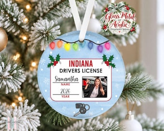May include: A round ornament featuring a light blue background with white snowflakes and a string of colorful Christmas lights. The design mimics an Indiana driver's license with the text Samantha, 2026, and a photo of a person holding car keys.