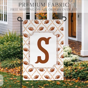 May include: A rectangular garden flag featuring a white background with a repeating pattern of brown footballs and orange autumn leaves. A large, centered brown letter S is enclosed within a decorative brown rectangular frame border.