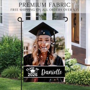 K&ouml;nnte beinhalten: Eine Gartenflagge zur Feier des Abschlusses mit einem Foto einer l&auml;chelnden Absolventin in Hut und Robe. Die Flagge enth&auml;lt den Text "Class of 2026" und den Namen "Danielle" auf einem schwarzen Banner. Oben auf der Flagge steht "Premium Fabric".