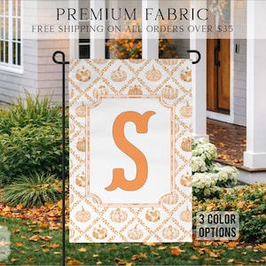 May include: A rectangular garden flag featuring a white background with a repeating orange pumpkin pattern arranged in a diamond grid. A large, centered orange letter S is enclosed within a decorative rectangular border on the flag's surface.