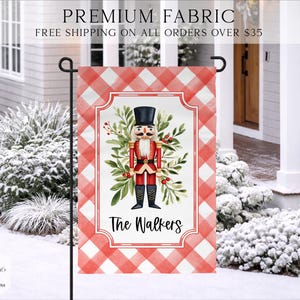 May include: A rectangular garden flag featuring a red and white buffalo plaid border. The center displays a nutcracker illustration surrounded by green foliage and red berries, with the text The Walkers printed in black script below it.