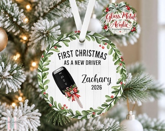 May include: A round white ornament with a wood plank texture, featuring a green mistletoe wreath border. It displays the text FIRST CHRISTMAS AS A NEW DRIVER, the name Zachary, and the year 2026, with a car key graphic adorned by a red bow.