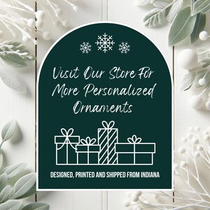 May include: A dark green arched sign with white text and snowflakes. The text reads "Visit Our Store For More Personalized Ornaments" above a graphic of wrapped gifts. The bottom text says "Designed, Printed and Shipped from Indiana".