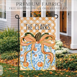 May include: A rectangular garden flag featuring a light orange and white gingham pattern at the top with the text THE CLARKS in black. Below, a large blue and white floral patterned pumpkin is adorned with a prominent orange ribbon bow.