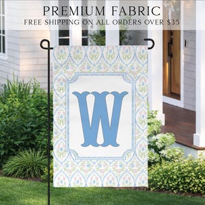 May include: A garden flag with a light blue letter "W" on a white background, framed by a decorative border. The flag features a floral pattern in blue, pink, and green. The words "PREMIUM FABRIC" are at the top.