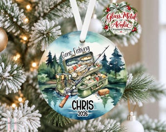 May include: A round hanging ornament featuring a watercolor illustration of a fishing tackle box, a fishing rod, and boots near a lake. The text reads Gone Fishing, CHRIS, and 2026. The ornament is suspended by a white ribbon against a blurred background.