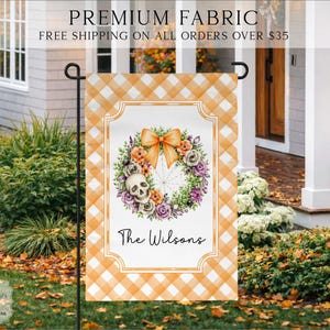May include: A rectangular garden flag featuring an orange and white buffalo check border. The center displays a white panel with a decorative frame, a floral wreath with a skull and orange bow, and the black cursive text The Wilsons.