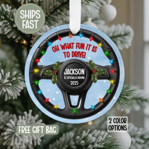 May include: A round Christmas ornament featuring a black steering wheel design with colorful lights. The text "OH WHAT FUN IT IS TO DRIVE!" is displayed, along with the name "JACKSON" and the year "2025".