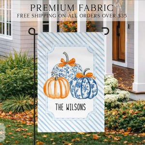 May include: A rectangular garden flag featuring a light blue and white diagonal striped background. The center displays three pumpkins, one solid orange and two with blue and white floral patterns, each topped with an orange bow. The text THE WILSONS is printed below.